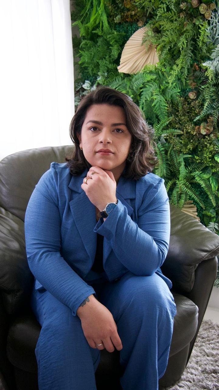 Stephanie Nascimento sentada, com a mão no queixo, de blazer azul, olhando para a câmera